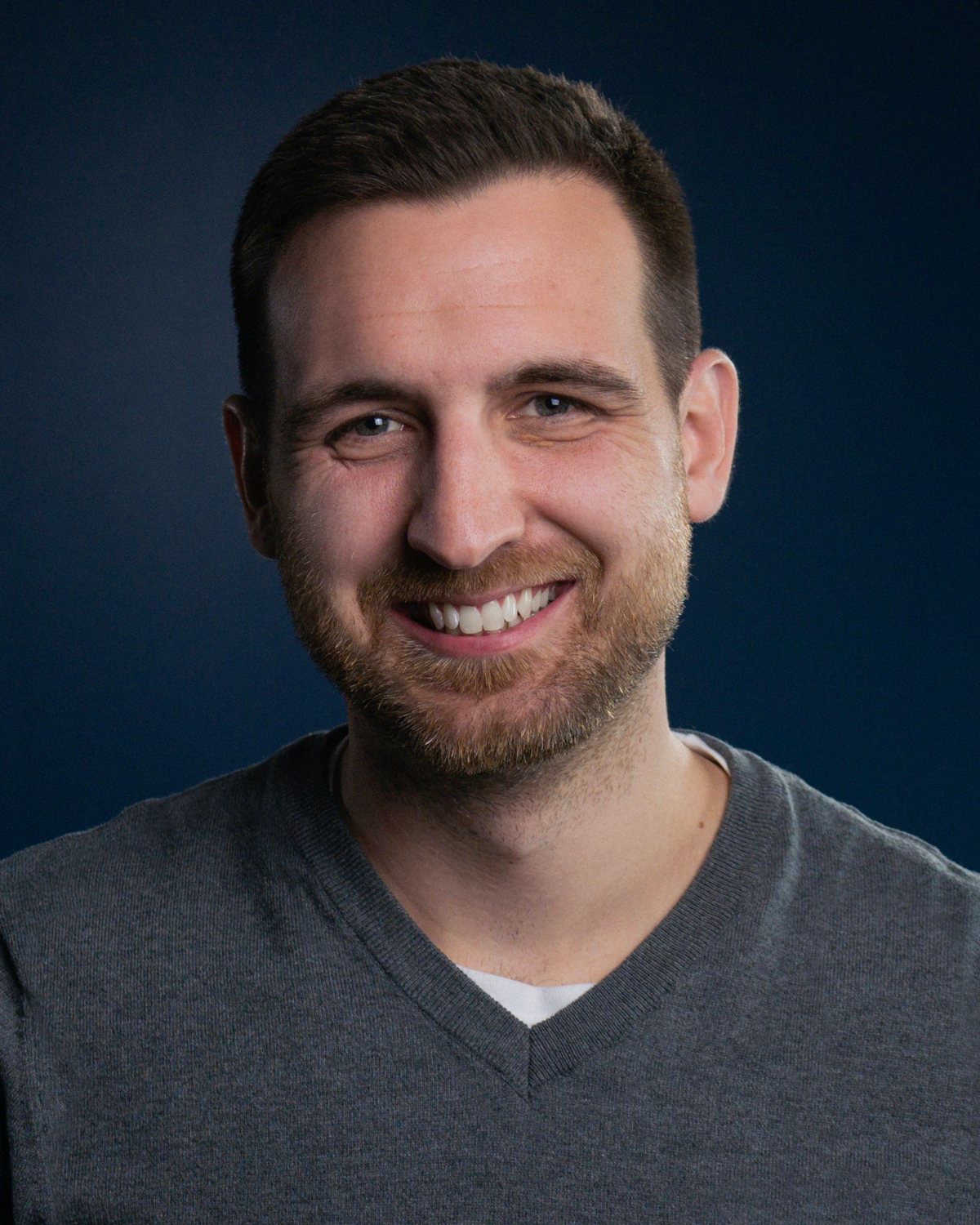 Spencer Martin, smiling, in a grey v-neck against a dark blue backdrop